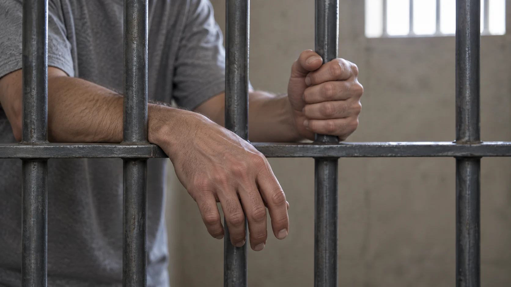 Hands reaching through and gripping steel detention cell bars, with no face visible